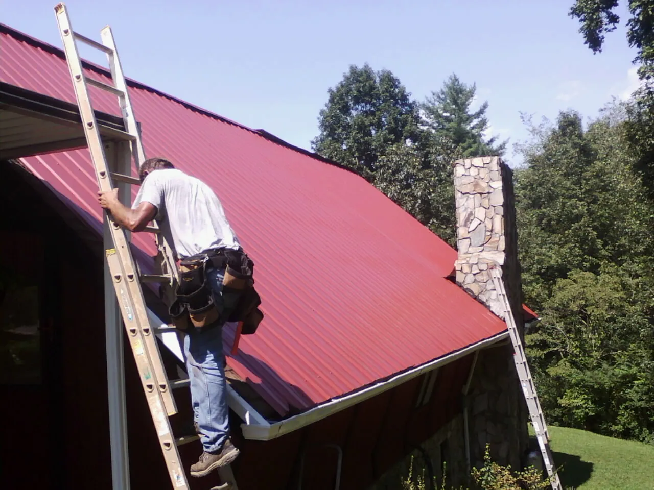 Roofer on a ladder beside a red metal roof and stone chimney — Soffit & Fascia Repair by Roofing Brothers in Patoka
